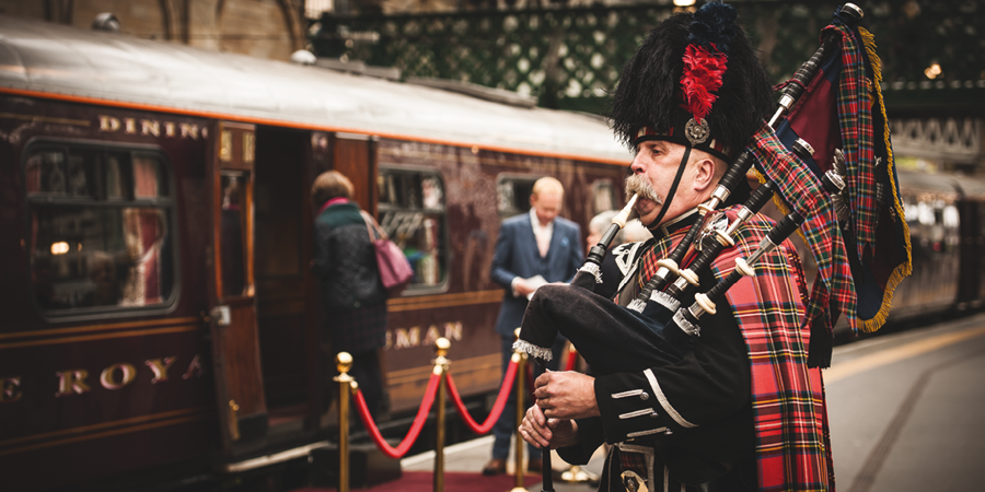 A man dressed in traditional Scottish military unform plays the bagpipes while passengers board the Royal Scotsman train.